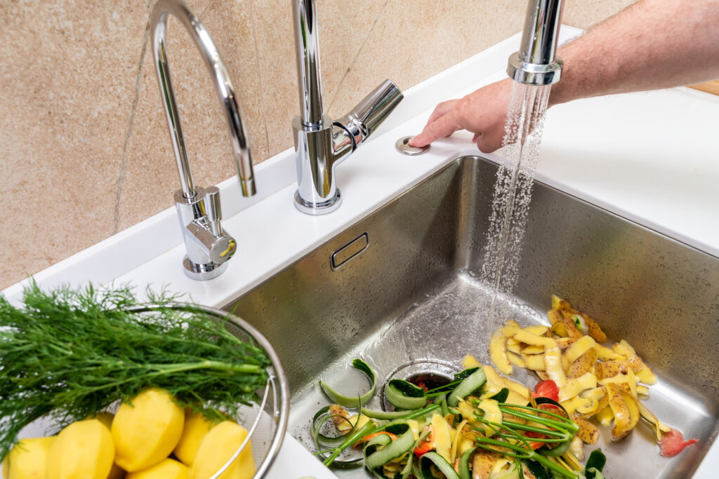 Person's hand turning on a garbage disposal to remove produce waste from a stainless steel kitchen sink.