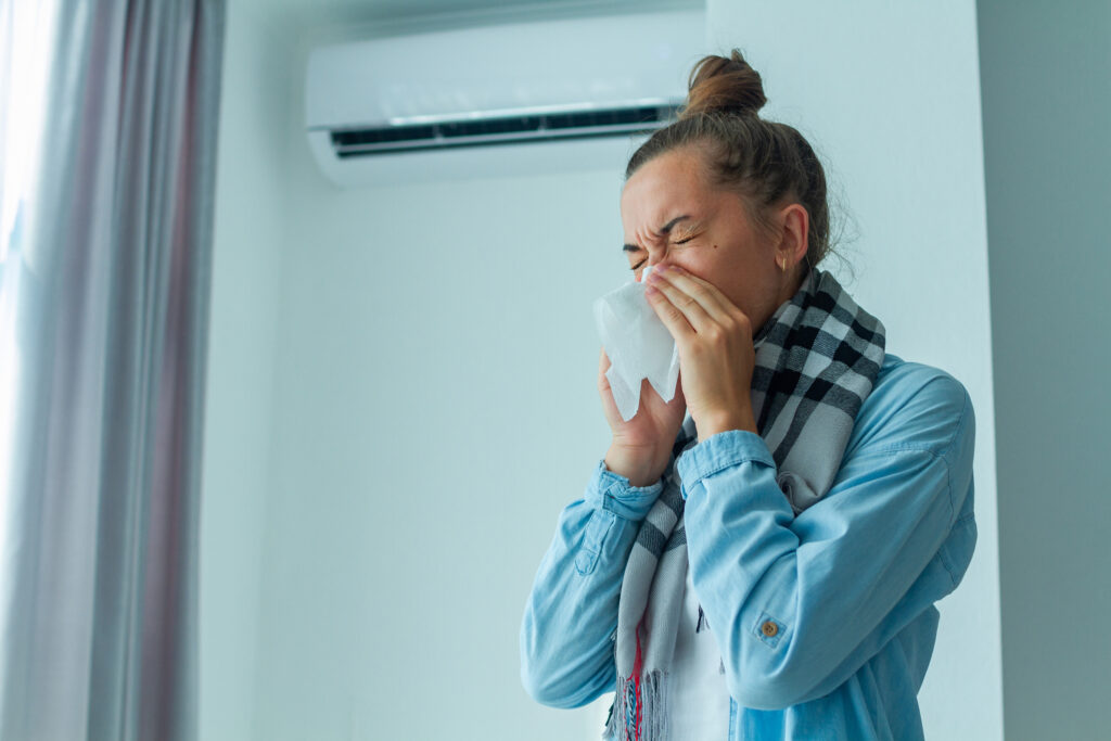 Woman sneezing from contaminated air, standing in front of dusty air system in her home.