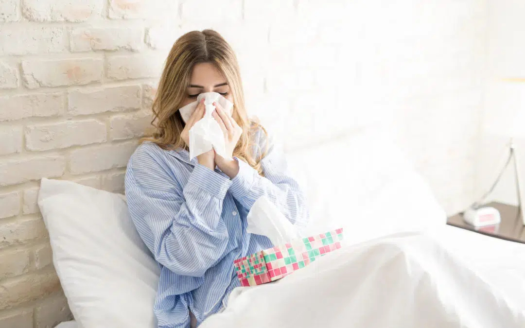 Woman sitting in bed with white sheets, white brick wall behind her. She has a tissue box on her lap and is blowing her nose.